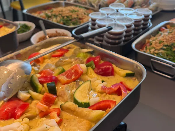 Un buffet installé sur un comptoir de bureau : un bac de curry aux légumes, deux plats de salade aux cacahuètes et des pots de sauce.
