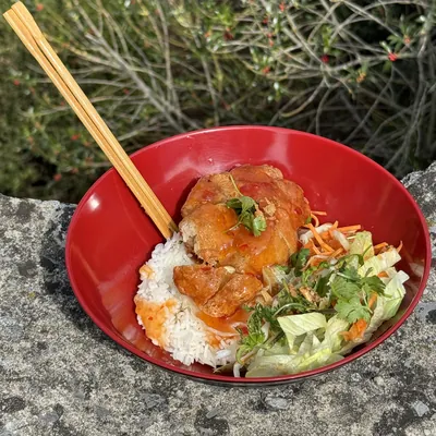 Un bol de riz blanc avec des morceaux de poulet en sauce, de la salade et des carottes râpées, des baguettes posées dans le bol.