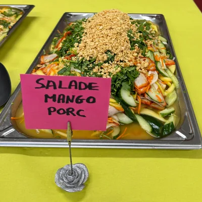 Un plat de salade mango sur la table du buffet, avec son étiquette écrite à la main : salade mango porc.